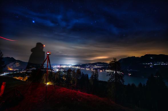 Nachtaufnahme einer weiten Aussicht auf eine beleuchtete Stadt am Ufer eines Sees, aufgenommen von einer Anhöhe. Im Vordergrund dominiert die dunkle Silhouette eines Fotografen, der mit einer Stirnlampe und einer Kamera auf einem Stativ auf einer rötlich beleuchteten, grasbewachsenen Fläche steht. Rote Lichtspuren der Stirnlampe ziehen sich durch den linken Bildbereich. Unterhalb der Anhöhe erstreckt sich ein Waldrand, dahinter die Lichter der Stadt, die sich am Ufer des Sees spiegeln. Der Nachthimmel ist tiefblau, mit Sternen und leichten Wolken, während der Horizont in warmen Orange- und Gelbtönen leuchtet. Die Szene vermittelt die Stimmung einer langen Nachtwache, in der vergebens auf Polarlichter gewartet wurde.
