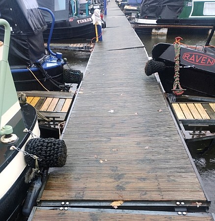 Photo along the length of a dock, made up of a chain of floating pontoons. The pontoons have a steel frame and a surface of wood planks. Each successive pontoon is rotated a few degrees to the left, so that the entire dock is curved.
The photo shows the sterns and bows of about ten narrowboats moored to smaller pontoons jutting off each side of the main dock. The narrowboat hulls are all black, but their superstructures have various colours—greens and blues—and black- or blue-fabric prams or cratches.