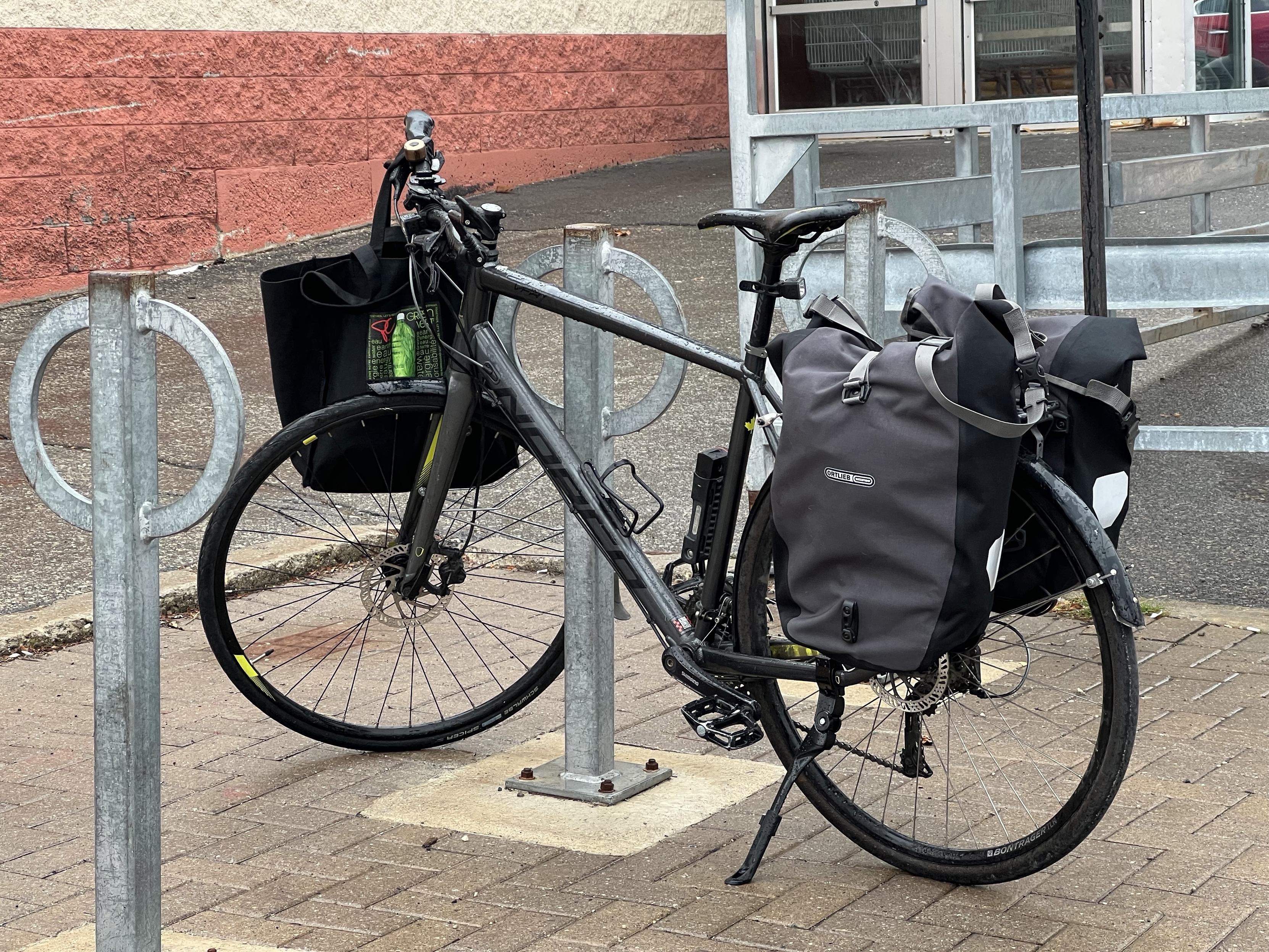 A side view of a bicycle parked next to a post-and-ring parking facility. The bike has two quite full panniers hanging over the rear wheels, and there’s a cloth bag hanging from the handlebars.