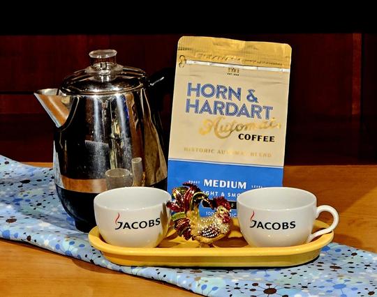 A variation on a tea tableau using coffee, instead. Upon a blue MidCentury atomic tablecloth there is a yellow Fiestaware oval tray holding two Jacobs coffee cups and a jeweled enameled rooster trinket box. Behind them are a shiny chrome MidCentury Modern coffee percolator and a blue and yellow bag of Horn and Hardart Automat coffee.