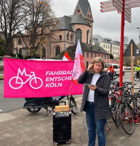 Fahrradbürgermeisterin Anna hält eine Rede zum notwendigen sicheren Radfahrstreifen auf der Rheinuferstraße. Im Hintergrund ein magentafarbenes Banner vom Fahrrad-Entscheid Köln und die Fahne der Kölner Kidical Mass.