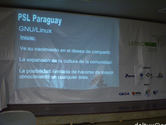 Lona branca cobre toda uma parede de sala de aula, havendo à esquerda a projeção de uma tela com o seguinte texto traduzido do espanhol: PSL Paraguai. GNU/Linux. Início: Vê seu nascimento no desejo de compartilhar. A expansão da cultura da comunidade. A possibilidade ilimitada de se transformar maior conhecimento em qualquer área. À direita, impressos na própria lona, estão logotipos do evento Latinoware 2008 e de organizadores e patrocinadores tais como Serpro, Celepar, PTI, Itaipu, Caixa, UOL e Dataprev.
