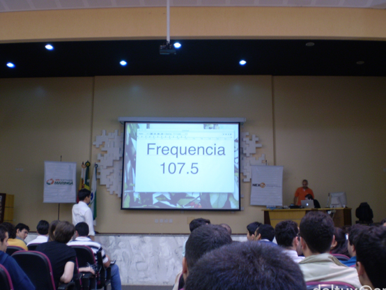 Do ponto de vista de uma pessoa sentada na plateia, cabeças observam a tela de uma projeção num palco em que se lê: Frequência 107.5. Há uma pessoa em pé diante do palco e outra atrás de uma escrivaninha sobre o palco.