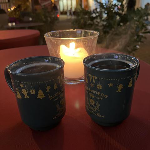 Zwei dampfende Glühweinbecher auf einem Tisch mit roter Tiechdecke. Dahinter eine Kerze in einem Glas. Das alles in einer Fußgängerzone.