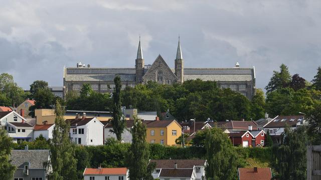 A photo of a large university building on the top of a hill. The hill has houses of different colors going up it with trees between them. The sky is cloudy.