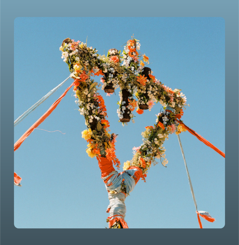 A large, three-pointed floral structure, made of green foliage and colorful flowers, is suspended in the air by orange and white ribbons. The sky behind it is clear and blue. The band name ‘THE DEVIL WEARS PRADA’ appears in small yellow uppercase letters along the top, and the album title ‘FLOWERS’ is written in bold yellow letters across the middle of the image.