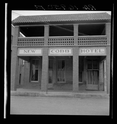 The black and white photograph depicts the Cobb Hotel with a two-story facade. The building is constructed of brick, featuring three prominent windows on each floor beneath an overhanging roofline supported by decorative beams. Above these openings are bold signs reading "NEW," "COBB HOTEL," and "HOTEL." Below this signage, there are wooden doors flanked by large glass-paneled entryways with mullions dividing the panes. The building appears to be unoccupied or closed as no people are visible in front of it nor does any sign indicate that it's open for business.

The photograph is marked "STERLING 1948" on a black line, which likely indicates either the photographer's watermark or a cataloging system used by Sterling. The image has an aged quality consistent with historical photographs from the mid-20th century. There are power lines running parallel above the building and faint markings or scratches visible in the negative at the top of the photograph.

The caption "Hotel in Memphis, Texas" suggests that this establishment is located in a town by that name within Texas, although no definitive reference to Memphis exists in known photographic archives from Dorothea Lange. The image may be part of an archive documenting historical sites or architectural styles during her time, possibly for sociological studies on the American South.

The website mentioned provides additional context and potentially more information about t [...]