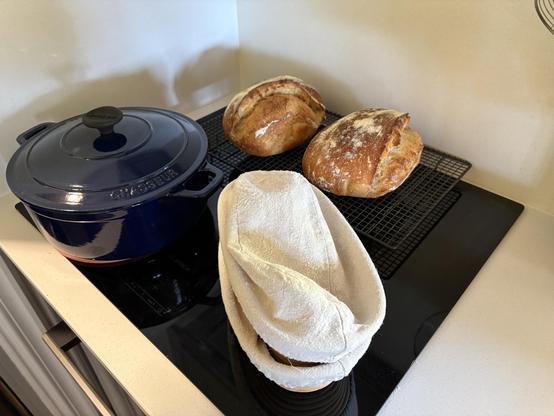 Two golden sourdough loaves sit cooling on a wire rack atop a kitchen bench. A blue cast iron pot and two bannetons are nearby.