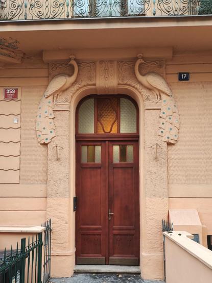 Art Nouveau doorway set into a peach-colored facade. Relief sculptures of peacocks with tail feathers are flanking the arched wooden door. Above the door there is a small arched window with warm-toned, decorative glass.
