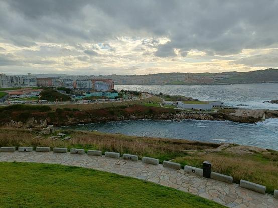 Imaxe da cidade da Coruña dende a Torre de Hércules. Vese a parte da cidade hacia as praias de Orzán e Riazor co paso marítimo así como o acuario. O ceo está nubrado, con algún claro ao fondo.