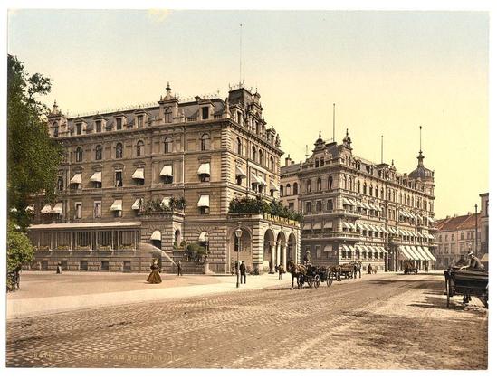 This image depicts a historic European street scene, likely in the late 19th to early 20th century. A grand building with multiple floors and ornate architectural details dominates the left side of the frame. The facade is adorned with numerous windows, some equipped with awnings or balconies, and decorative elements such as finials at the roofline.

In front of this imposing structure, a woman in period attire sprays water from a large yellow watering can onto what appears to be cobblestone pavement. She stands out due to her colorful dress against the muted tones of the architecture. The street is bustling with activity: people walking on footpaths or crossing streets, and horse-drawn carriages suggest an era before automobiles became commonplace.

The right side features another building with a similar architectural style, though less ornate in comparison. Street lamps line both sides of the road, adding to the historical ambiance. A bench sits empty by the curb, inviting passersby to rest. The overall impression is one of daily life and commerce within an urban European setting from that period.

The image's sepia-toned coloration hints at its age, with a vintage feel consistent with early photographic processes such as albumen prints or hand-tinting on cartes-de-visite. This particular photograph was taken in Bremen, Germany according to the caption provided and is part of the Loener Collection (https://images.loene [...]