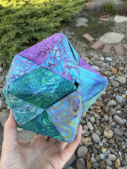 Purple greens and turquoise fabrics, folded into an octahedra with rocks in the background this angle shows a non-symmetrical view.