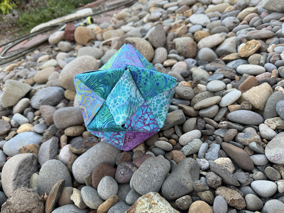 A fabric octahedron with purple, turquoise and green fabric on river rock with one of the points visible as a triangle.