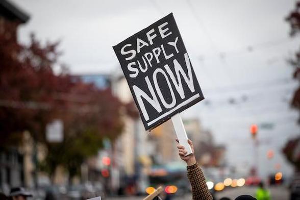 A person holds a black protest sign reading “SAFE SUPPLY NOW” in bold white letters. The sign is raised above a crowd on a city street, with blurred buildings, trees, and traffic lights in the background on an overcast day.