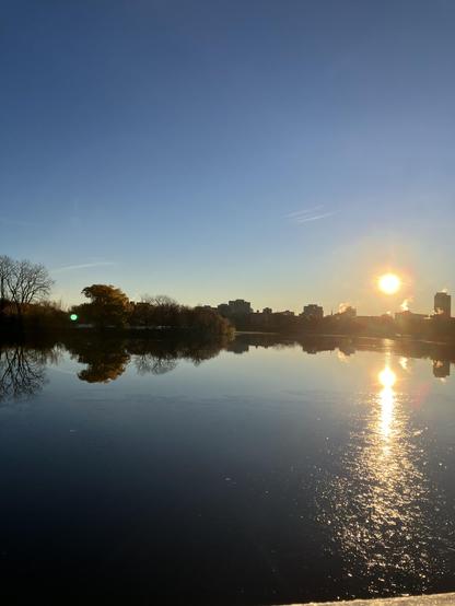 Sunrise over calm water with blue sky reflecting along with the flowing sun. Trees and buildings mid frame have a gentle glow behind them