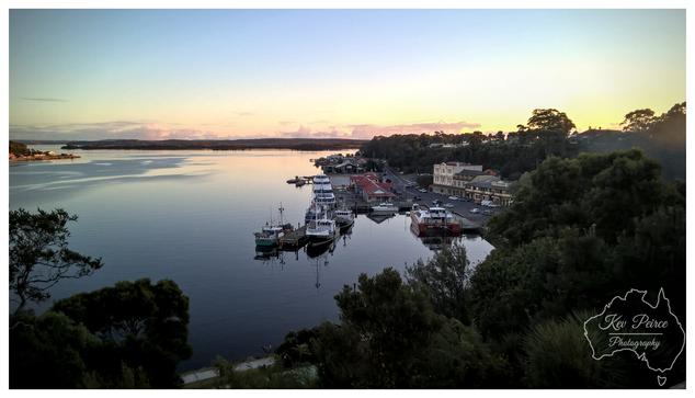 A tranquil, high angle view of the Strahan waterfront at sunset, looking across Macquarie Harbour. The sky features a soft, pale blue transitioning to golden pink light just above the distant tree lined horizon.  The water is still and dark, reflecting the colours of the sky. In the middle ground, a cluster of fishing boats and small commercial vessels are tied up at a pier, adjacent to historic town buildings nestled among dense greenery.  Dark foliage frames the bottom and right side of the photograph.