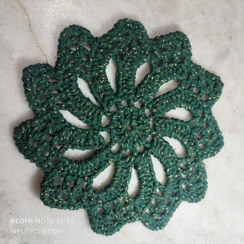 Descanso de prato, descanso de panela ou pequeno forro de crochê. O fio é barbante verde escuro enfeitado com fio dourado. A peça tem um formato de flor estrelada, com onze pétalas.