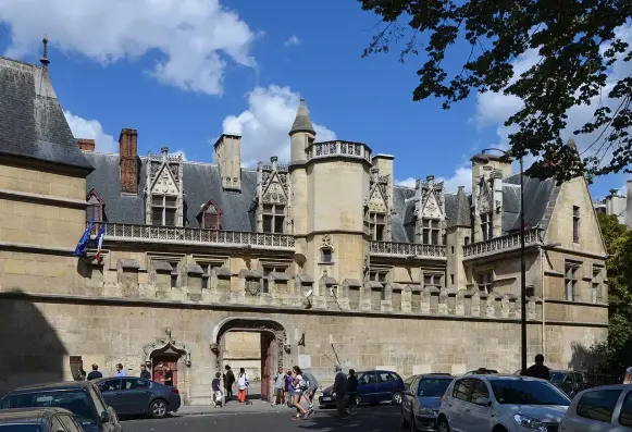 The image depicts a historical building, likely a château or large estate, characterized by its distinct architectural features such as turrets, intricate stonework, and decorative elements. The scene includes people walking in front of the structure and several parked cars. The sky is partly cloudy, enhancing the overall ambiance of the location.

Image Credits: Wikimedia / Pline / CC BY-SA 3.0
