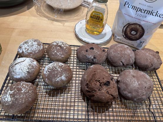 Wire baking rack with five lighter brown and five darker brown rolls. Ingredients visible in back: honey jar on saucer and bag of King Arthur pumpernickel flour.