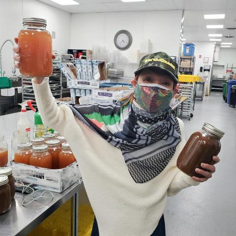 Me, wearing a camouflage hat, sequined mask over N95 mask & a Palestine scarf, holding a large glass of jar fig jam in my left hand & a jar of apple juice, above my head with right hand. I am standing in a commercial kitchen with jars of apple juice on a stainless steel work surface in the foreground, boxes of jar lids labeled "BERNARDIN" behind them, and a clock on the wall. The hat says "AnarchoSocialist" in yellow.