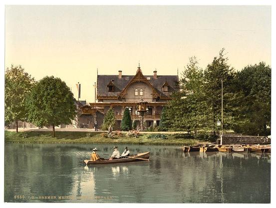 The image depicts a serene lakeside scene with an old-fashioned, multi-story building as the central focus. The architecture of the structure suggests it may be from around late 19th to early 20th century Europe. It has steeply pitched gable roofs and ornate wooden detailing typical of that era's residential design.

In front of this house is a calm lake with two individuals in a rowboat, engaging in what appears to be leisure paddling. Both are wearing hats indicative of the fashion from around 1890-1900s Western Europe, suggesting they might be out for recreation or social outing at that time.

The surroundings include lush greenery and trees on either side of the lake's bank, enhancing the tranquil atmosphere. Several small boats docked by a wooden jetty are also visible, reinforcing the recreational nature of this setting. The overall color palette is muted with earthy tones dominating, giving it an aged or vintage feel consistent with historical photographic methods.

The text at the bottom left indicates that this image has been catalogued as "6880 P.Z. BREMEN. MEIHL'S HORTSPARK," suggesting a connection to Bremen and possibly Hortspark—a park in the city of Bremerhaven, Germany—as referenced by Loener's archive (Views of Germany). The photochrom print suggests it was created using a process known as Photochrom printing that produced colored images from black-and-white photographs.