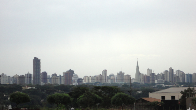 Fotografia diurna com um panorama de edifícios do centro da cidade de Maringá, à distância de alguns poucos quilômetros, após muitas árvores verdes que integram sua urbanização.