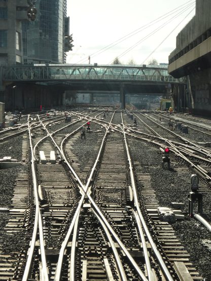 Ferrovia com cerca de dez pares de trilhos que seguem paralelos em direção à vista da fotografia, com muitos autos de mudança de via, e vários pequenos semáforos que estão vermelhos. A ferrovia está em meio a edifícios e, ao fundo, há viadutos.