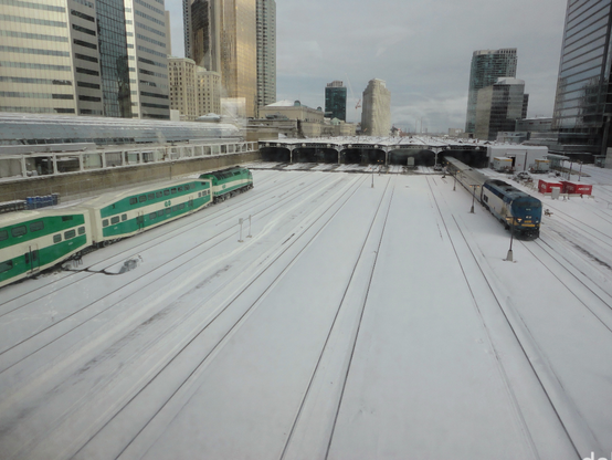 Ferrovia com uma composição chegando e outra partindo de uma estação vista a distância, com muitas vias paralelas em meio ao terreno coberto por neve. O trem que chega, em verde e branco, tem carros de dois andares com marca GO, do Governo de Ontario. Já o que parte tem uma locomotiva azul com marca da empresa Via Rail. O céu está nublado e se avistam edifícios altos espelhados aos lados.