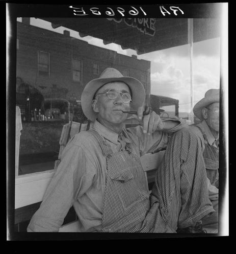 The photograph is a black and white image depicting two men seated inside what appears to be an establishment, possibly a diner or store. The primary subject in the foreground wears glasses, a wide-brimmed hat, suspenders with striped overalls tucked into them, and has his hand resting on a bench seat while holding a pipe close to his mouth as if smoking it. His facial expression is contented and relaxed.

The second man sits beside him, slightly out of focus in the background, wearing a similar wide-brimmed hat indicative of rural or agricultural life, suggesting both individuals are likely farmers given their attire that's practical for manual labor such as farming. Behind them, through what seems to be a window reflecting on another surface, is an establishment with signage partially visible and some interior details like shelves containing bottles.

The image exudes a sense of casual reprieve from work or travel, capturing a moment in the lives of Dust Bowl farmers during their time spent away from fields for social interaction. The setting implies it might be a temporary stop on their journey across Texas as part of migratory farming practices associated with the Great Depression and its aftermath.

Additional context is provided through external resources indicating this photo's connection to "Dust bowl Farmers of West Texas," hinting at economic hardship, movement due to droughts in agriculture communities, and so [...]