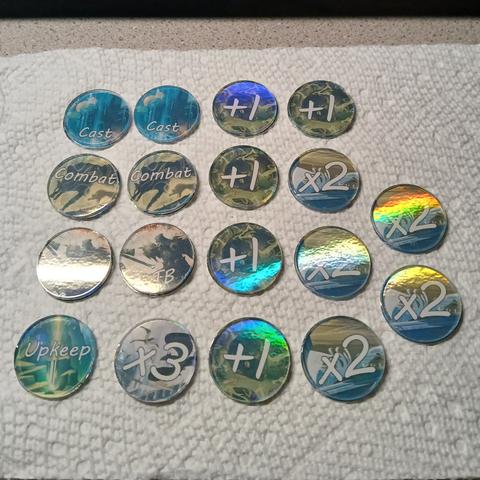 Photo of 18 round tokens: two say Cast, two Combat, two ETB, one Upkeep, one x3, five +1, and five x2. The tokens have various MTG card art as the background and the text is in white. Each token has rainbow foil and is topped with a clear acrylic disc.
