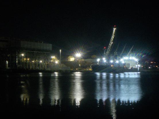A night time shot of a cargo ship at a dock, with cranes above it and a structure to the left.  Lotta flare around the lights