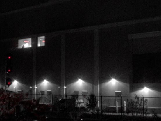 Black and white image of the side of a building.  There is a railroad signal in the left foreground with red lights, and a window with a human silhouette in it in the upper left