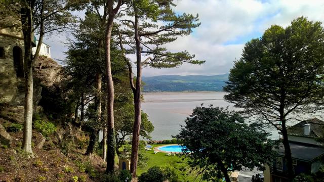 Trees on a rocky hillside overlook a calm estuary with low tide exposing sandbanks, while green hills rise in the distance under a partly cloudy sky. A small circular swimming pool sits on a neatly kept lawn near the water’s edge, surrounded by lounge chairs and bordered by shrubs and flowers. To the left, part of a stone building clings to the slope, and to the right, rooftops of other structures appear between the trees. The scene combines natural woodland, open water, and landscaped grounds in a quiet, scenic setting.