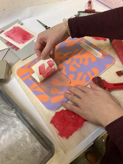 Gellipainannan ensikokeilu: värikkään tarjottimen ympärillä on maaliteloja, maalia ja erilaisia kortti- ja paperipohjia kokeiluihin. Tarjottimen päällä Kaislan kädet paljastavat gellipainannan tuloksen, vetäen geelimäiseltä levyltä siistin monoprintin, jossa näkyvät selvästi punaisella maalilla vedetyt siveltimenjäljet.