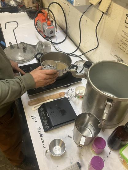 Kaikenlaista keittiökemian roinaa valkoisella pöydällä, ja mies työskentelemässä niiden äärellä. Induktiolieden levyllä kattilassa on agar-agaria, ja siihen ollaan kaatamassa lasista glyseriiniä. Kauempana näkyy lämpömittauksen laitteita ja niiden johtoja sekä painekattilan kompressori. Etualalla painekattila sekä mittavälineitä, vaaka, vispilä, lasta, ruskea pullo, ruisku sekä agar-agar-purkkeja.