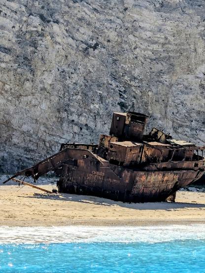 Ein älteres Schiffswrack liegt vor einer steilen Felswand auf einem Sandstrand. Das Meer davor ist türkisblau. Das Metall des Schiffswracks ist durch und durch verrostet.