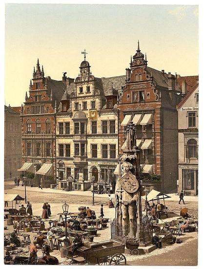 This image depicts a vibrant street scene in what appears to be an old European town, possibly Bremen given the caption. The architecture is characterized by ornate facades with gabled roofs and decorative brickwork typical of historical German towns during this period. A prominent feature on the left side includes a statue or monument placed at the forefront. In front of it, there's an outdoor market bustling with people engaging in commerce. Vendors are seated around tables laden with goods such as fruits and vegetables, suggesting a marketplace atmosphere.

The street is lined with pedestrians walking by, some carrying umbrellas, indicating variable weather conditions typical for that era. The buildings have multiple floors, displaying various shop fronts at the ground level, hinting at mixed-use architecture common in historical towns where residential spaces often doubled as workshops or stores.

Street lamps and what appear to be horse-drawn carriages add a sense of antiquity to the scene. Signage on some building entrances provides hints about businesses housed within these structures, though not all is legible from this distance. The overall palette consists of muted earth tones with splashes of color coming from clothing and market stalls.

The vintage look suggests that this image was taken using photochrom printing techniques popular in Europe during the late 19th to early 20th centuries. Photochrom prints wer [...]