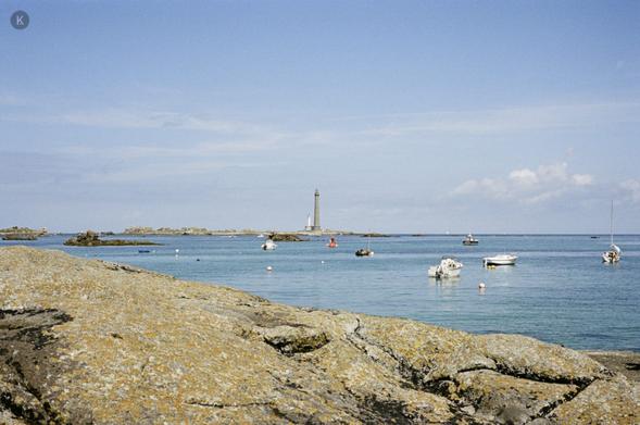 Ruhige Küstenlandschaft: Im Vordergrund helle Felsen, dahinter türkisblaues Meer mit kleinen weißen Booten und Bojen; am Horizont ragt ein schlanker Leuchtturm unter blauem Himmel mit wenigen Wolken auf.