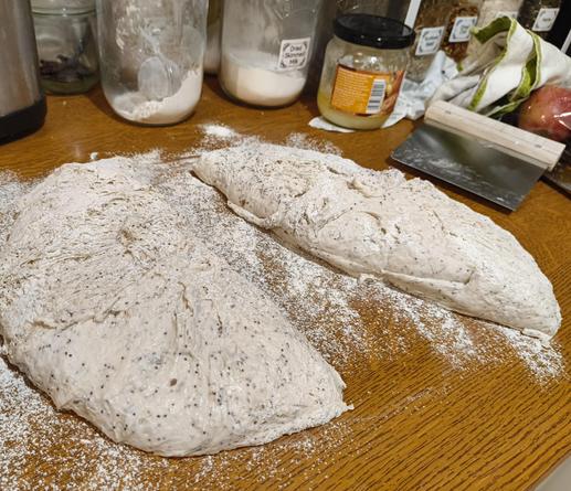 A floured bread dough on a worktop, roughly split into two pieces. You can see grains in the dough and tell that it's a wet mix by the way it spread out on the counter.