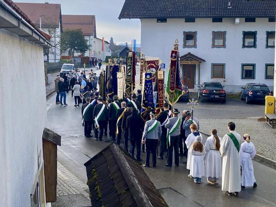 Foto, das von etwas erhöhter Warte aus einen dörflichen Straßenzug zeigt, auf dem sich ein Prozessionszug aufgestellt hat. Der Zug ist von hinten gesehen. Den Haupttrupp bildet eine Gruppe von Männern mit grün-weißen Schärpen, die ziemlich viele schwer hängende, bestickte Vereinsfahnen tragen. Dahinter, also näher an der Kamera haben sich vier Kinder vom Altardienst mit Vortragskreuz und ein Geistlicher mit grüner Schärpe aufgestellt, sie tragen weiße Gewänder. Weiter hinten sind weitere Leute, und auch das Ziel des Zuges ist zu erraten: ein Kriegederdenkmal, neben dem rechts uns links zwei Fahnen aufgezogen sind: die Deutschlandfahne und die Bayernfahne. Vor einem Haus rechts der Straße sind parkende Autos und ein gelber Briefkasten zu sehen.