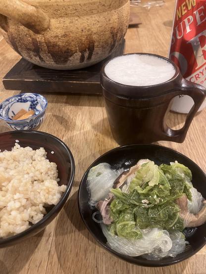 A photo of a meal featuring a clay pot, a black bowl with glass noodles and leafy greens, a bowl of brown rice, and a frothy beverage in a wooden mug. A small blue dish containing crackers is also visible