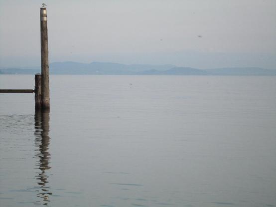 Le lac Majeur du côté italien, légèrement brumeux avec sur la gauche un ponton de bois et à l'horizon les Alpes bleutées.