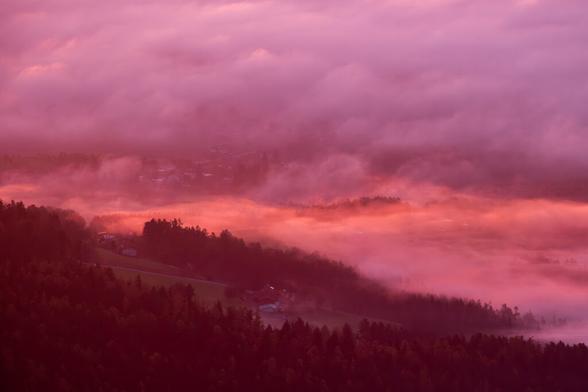 Blick von oben auf ein Tal. Im Vordergrund ein herbstlicher Wald. Nach hinten abwechselnd Wiesen mit Häusern und Wälder. Im mittleren Teil des Bildes beginnt leichter Bodennebel, der sich nach hinten auftürmt und zu einer dicken Hochnebeldecke wird. Teilweise ist der Nebel von der einstrahlenden Sonne erhellt. Das Bild hat einen morgendlichen lila Farbton.