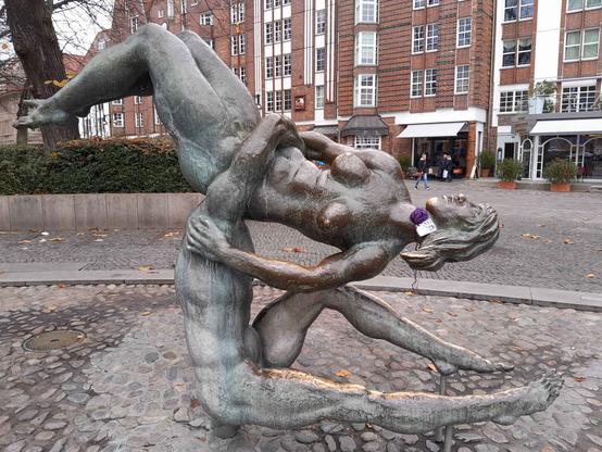 A bronze sculture of a man and a woman. Both are naked the man is sitting and balances the woman with one arm on his shoulder. The woman is hold in a horizontal position with her face looking up. Both are part of a fountain.
A purple crochet rose is tied around her neck. With a label attached. The label says Stop Gender-Based Violence #rosesagainstviolence