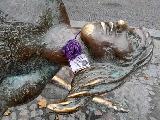 A close up of the sculpture showing the womans head with the attached crochet rose.
The rose has a label with a hand print saying. "Stop genderbased violence!" #rosesagainstviolence
The woman's chin, lips and nose have noticeably lighter bronze colour from touching.