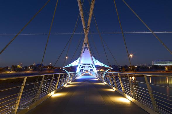This is an abstract colour sunset photo in landscape format looking along a pedestrian bridge. Tempe, Arizona (2012).

The camera is aligned with the centre of a modern pedestrian bridge, with footlights lighting the ground, spaced every few metres. Along either side are two metal railings angled inwards about thirty degrees from the vertical. These railings converge together with increasing distance so as to nearly touch at the end of the bridge, perhaps a hundred metres away. About ten metres away are two white tubular, near vertical frames forming an 'X' with stout rods descending downwards to the bridge in the fashion of a cable-stayed suspension bridge. Just beyond the support is an illuminated awning stretched out in a sharp, pointed triangle. A second 'X' frame can just be made out in the distance. On the right side of the bridge is a still, body of water. On the left side - darkness. Above our bridge is a clear and cloudless blue sky, slightly orange along the ground but rapidly graduating into blue and becoming darker towards the top of the image. About two-thirds up in the image is a straight, bright line stretching across the frame from left to right, with pairs of white dots of lights evenly spaced above the line, while on the line are occasional red dots of light. 

The bridge is the Tempe Dam Pedestrian & Bike Bridge, with the Tempe Town Lake on the right of the photo. The line of light is from an aircraft landing a Phoenix airport.