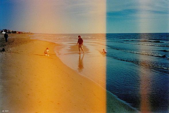 Farbfoto, aufgenommen am Strand. Rechts das Mittelmeer mit ein paar Wellen, in Bildmitte, im Wasser stehend, ein Mann. Links und rechts von ihm kleine Kinder. Fotografiert mit einer alten Olympus-Filmkamera, die einen Defekt hatte, weshalb etwas mehr als die linke Bildhälfte in oranges Licht getaucht ist.