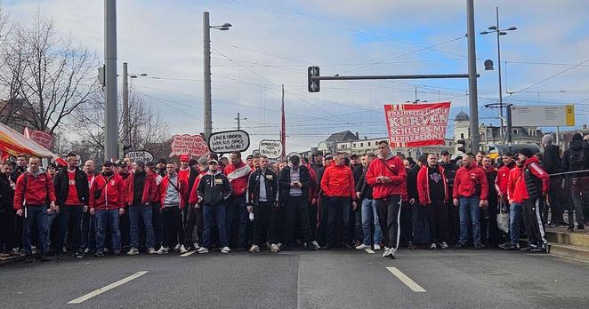 Eine große Gruppe von Personen steht auf einer breiten Straße, die von einem Gehsteig gesäumt ist. Die meisten Personen tragen rote Jacken und dunkle Hosen. Einige Personen halten Banner und Schilder hoch. Im Hintergrund sind Bäume und Gebäude zu sehen. Ein Verkehrslicht ist über der Straße zu sehen. Auf einem der Banner steht: "FREIHEIT FÜR DIE KURVEN".