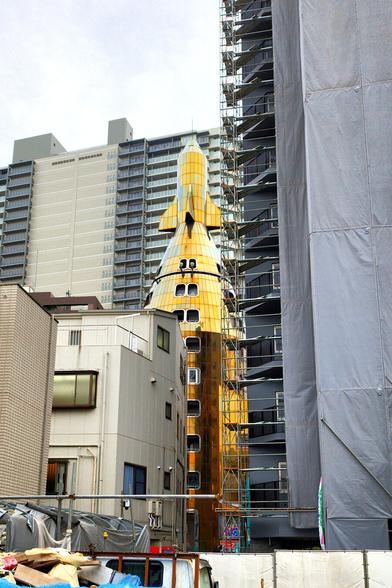 We are looking over the top of a truck cut off at the bottom and past a high rise building under construction on the right at the center, where a building shaped like a rocket stands in the middle of it all. It's 5-6 storeys tall. The building is a round booster rocket and a miniature space shuttle is at the top. It's coated in golden metal plating. Both the building under construction and some low nondescript buildings are blocking the view a bit. There is another big blocky building in the background and just a bit of hazy sky in the upper right hand corner.