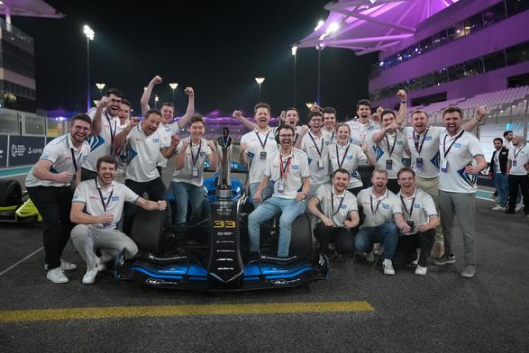 A large group of TUM Autonomous Motorsport team members celebrates around their blue race car on the Yas Marina Circuit at night. They are smiling, cheering, and posing with the winning trophy, with the illuminated grandstands in the background.